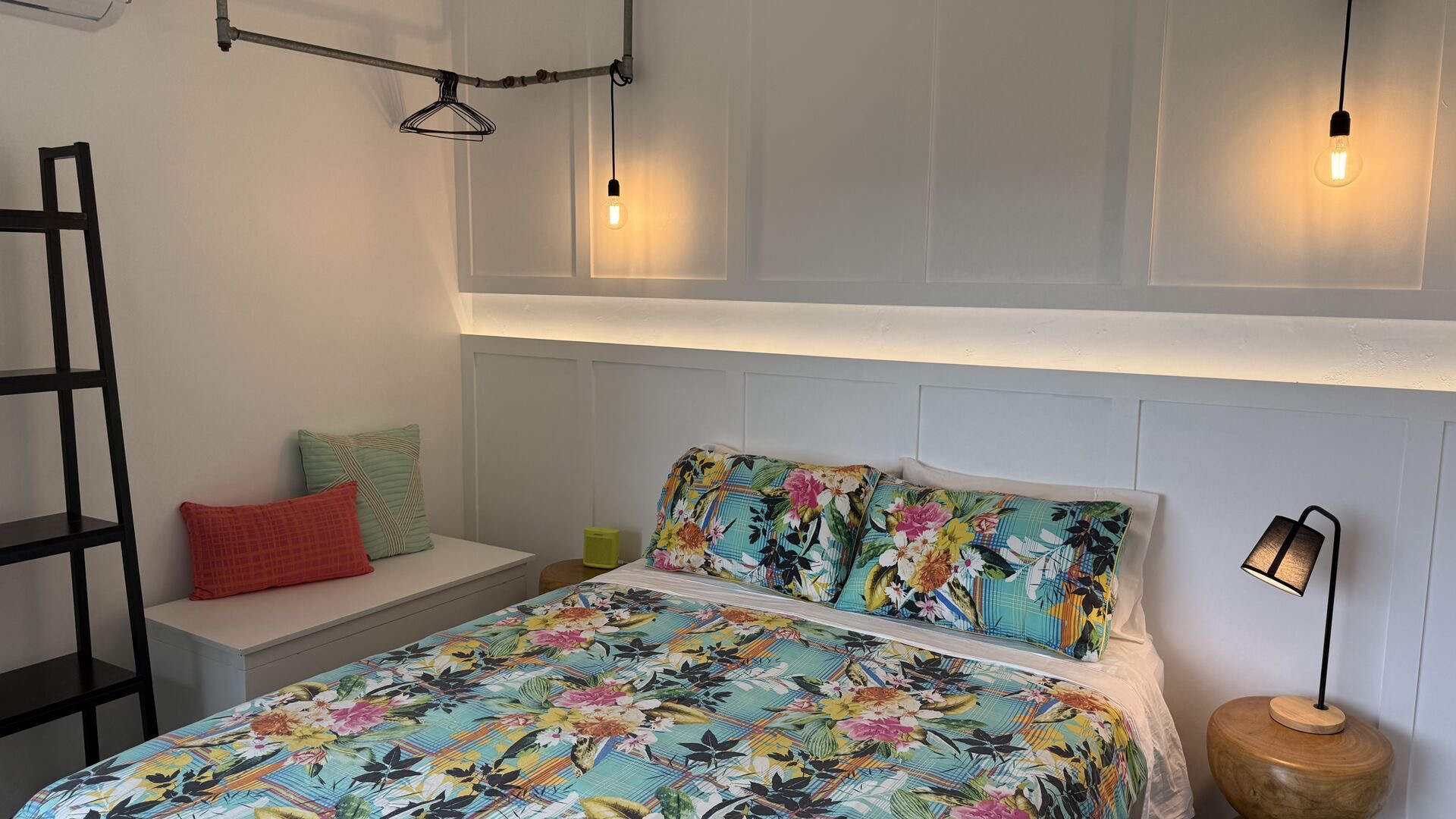 The bedroom with bright tropical floral bedding, a black ladder shelf, a turned-timber side table and warm pendant lighting.
