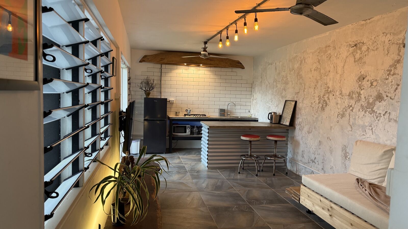The living room with doors opening onto the lawn and lagoon beyond, a live-edge media bench, Edison-bulb track lighting and a chalkboard welcome sign.